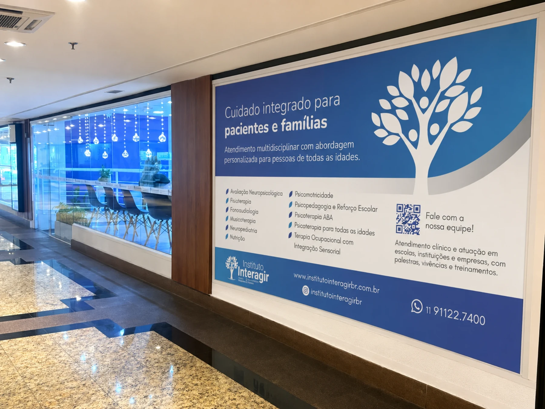 Painel de comunicação visual na entrada da clínica, com o símbolo em branco sobre azul, a chamada “Cuidado integrado para pacientes e famílias” e a lista de especialidades.